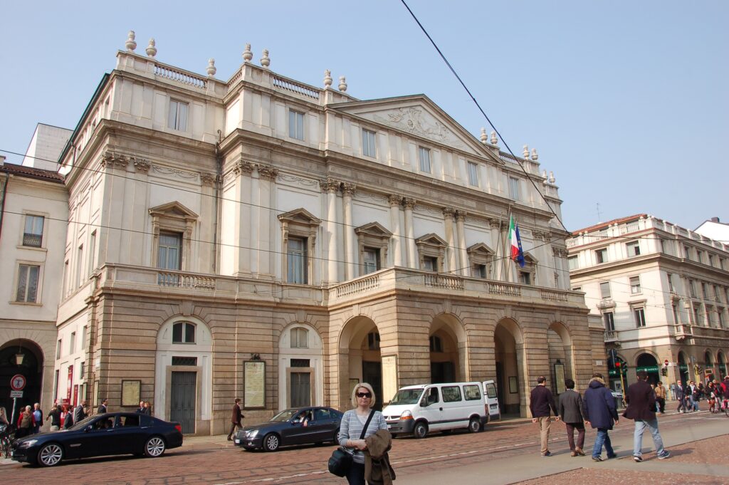 Navigli csatorna kivilágított házakkal és esti hangulattal Milánóban és Teatro alla Scala épülete nappali fényben, előtérben Leonardo szobrával